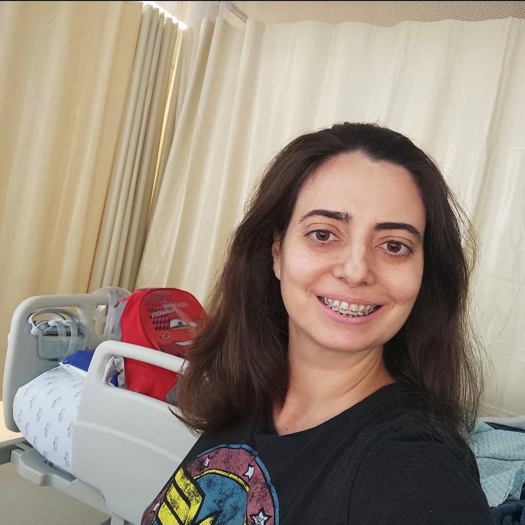 Sabrina Malichésqui no quarto do hospital, sorrindo, com a camiseta da Mulher Maravilha e a mochila do McQueen ao fundo